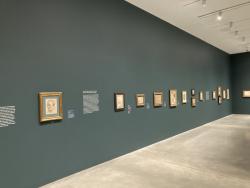 Paintings in gold and brown frames hang on an olive green wall of the Berkeley Art Museum Pacific Film Archive exhibit. 