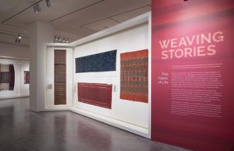 display of handmade textiles on a white gallery wall next to a red wall with the title 'Weaving Stories' and the description under.