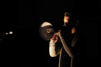 Ken Ueno speaks into a megaphone in a dark room.