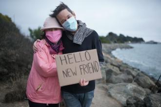 An older women in a pink jacket and a younger male in a black sweater hug each other near the marina while holding a cardboard sign that says Hello New Friend