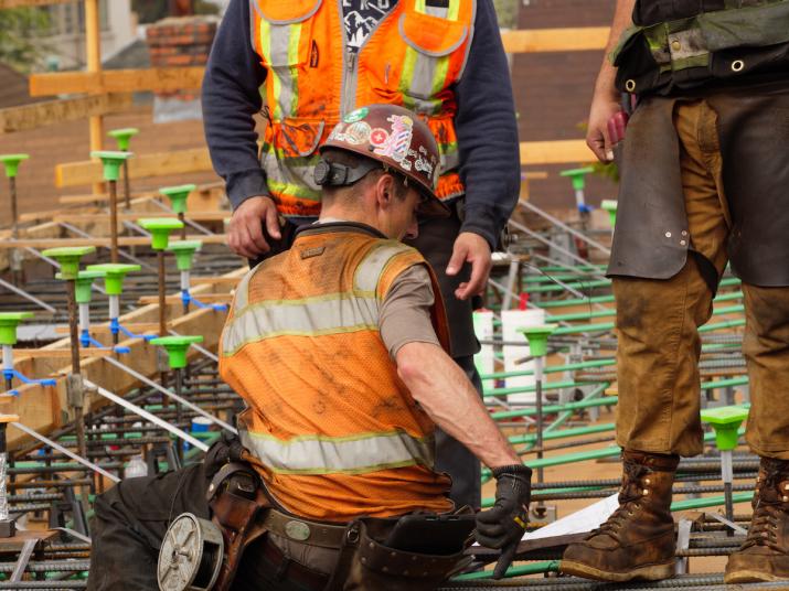 One construction worker in a brown hardhat covered with stickers and an orange and neon yellow vest sits down while two other construction workers stand around him.