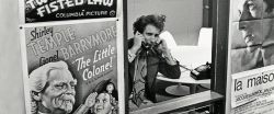 In this black and white image, a man talks on an old telephone through an open window. Old posters surround the open window.