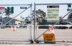 a closed fence with the one orange sign on the floor reading "vaccine lane," and one yellow sign on the fence reading "Restricted Area no thru traffic"
